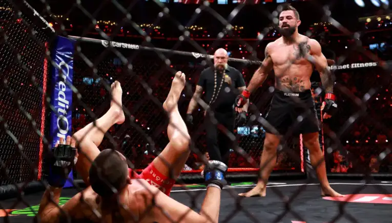 Muscular MMA fighter stands inside the fenced octagon as an opponent lies on the mat with legs up, referee watching in the background.