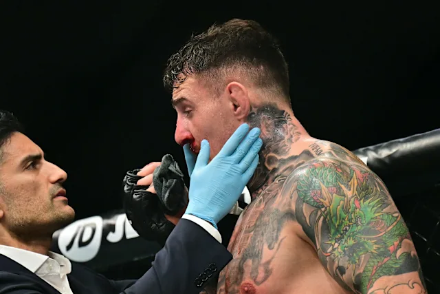 Boxer with colorful tattoos receives care from a trainer wearing blue gloves in a boxing ring corner.
