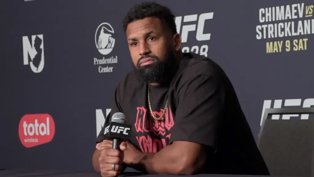 UFC fighter with a beard sitting at a press conference, holding a microphone.