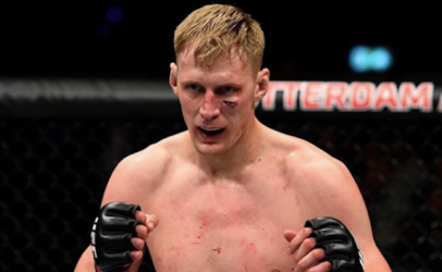 Male mixed martial artist inside a cage, fists up in black gloves, focused expression and visible minor bruises on face and torso.