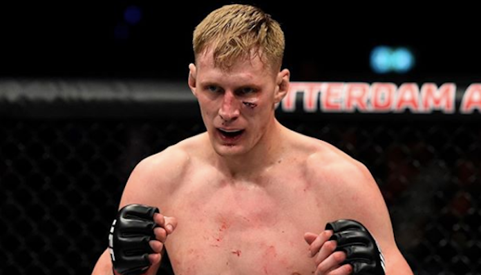 Male mixed martial artist inside a cage, fists up in black gloves, focused expression and visible minor bruises on face and torso.