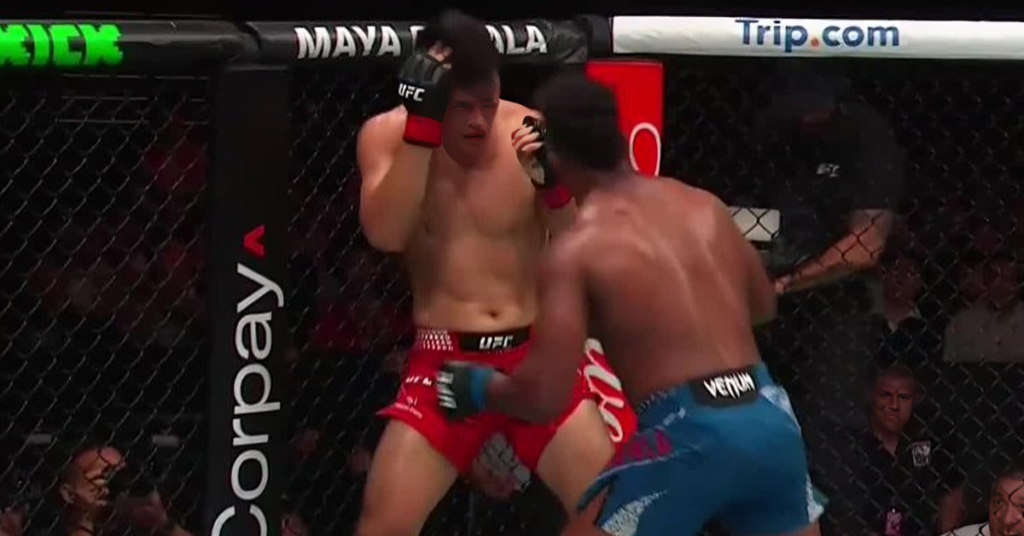 Two male UFC fighters exchange punches inside the octagon, one in red shorts and one in blue shorts, surrounded by a cage and sponsor banners.
