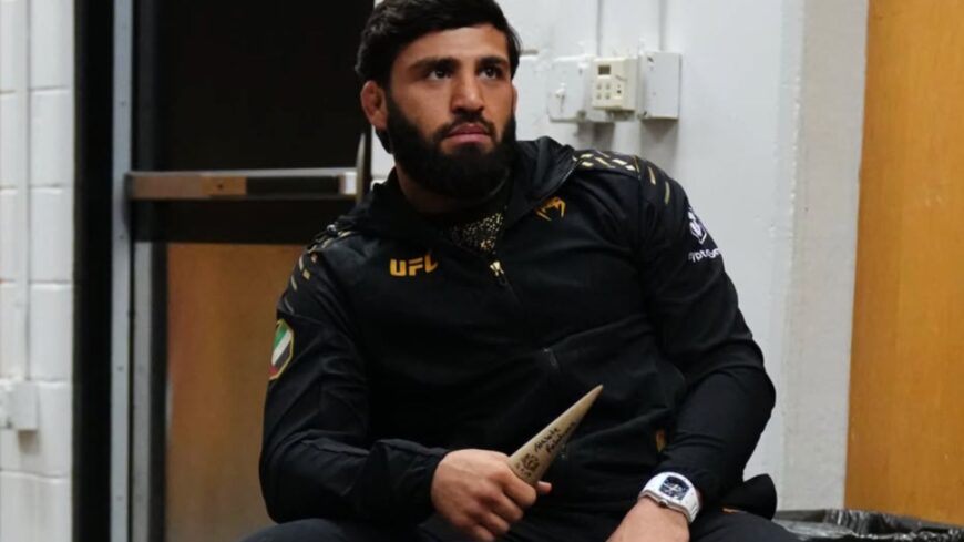 Bearded man in a black UFC jacket sitting in a hallway, holding a ceremonial dagger.