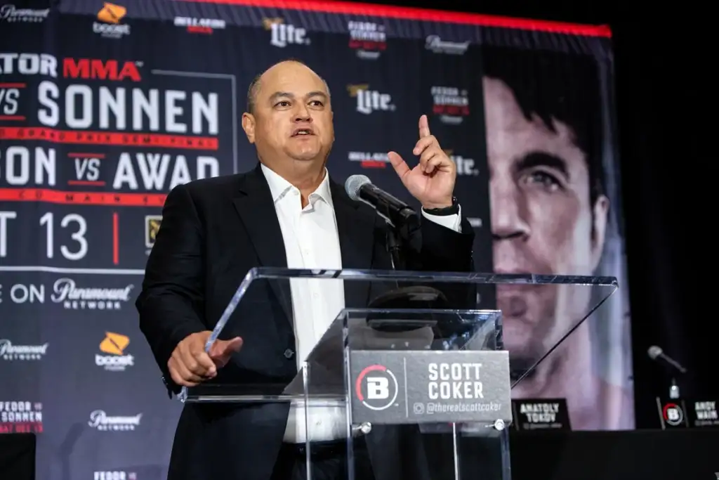 Scott Coker speaks at a press conference from a clear acrylic podium, gesturing with his right hand against a dark MMA event backdrop.
