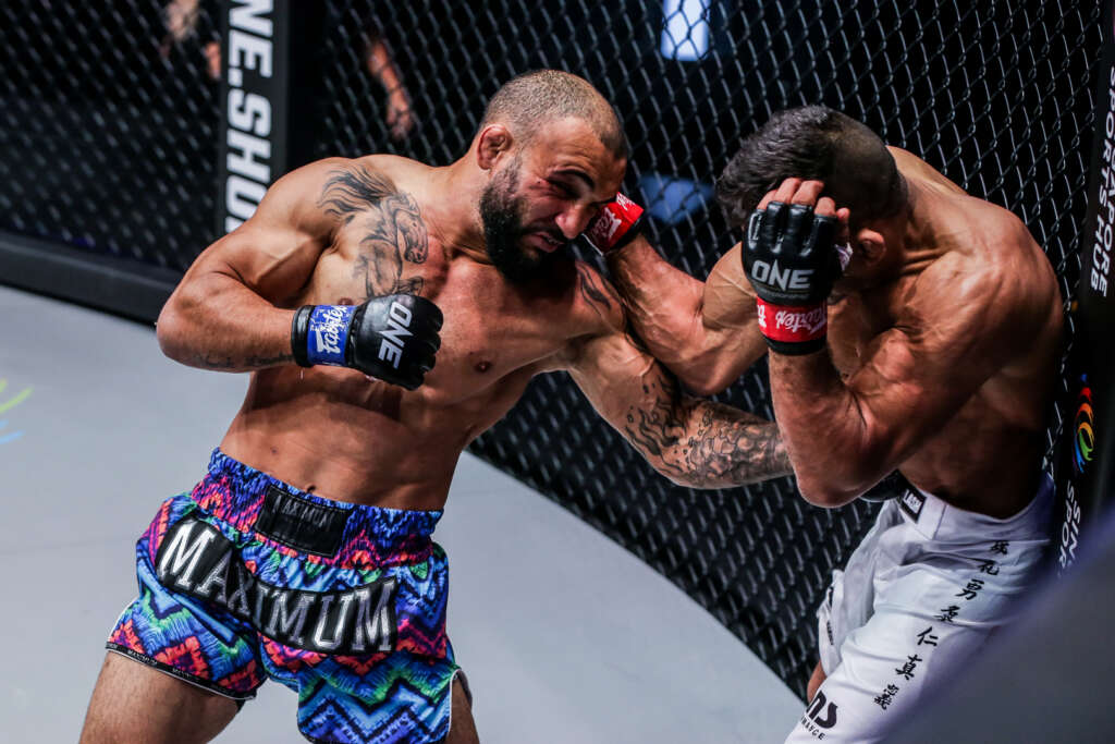 Two MMA fighters clash inside a chain-link cage; left fighter throws a punch as the right defends with his forearms up in a tight guard.