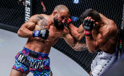 Two MMA fighters clash inside a chain-link cage; left fighter throws a punch as the right defends with his forearms up in a tight guard.