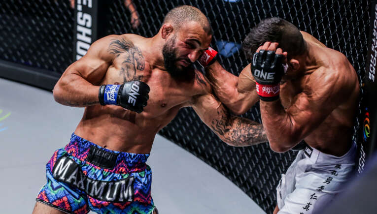 Two MMA fighters clash inside a chain-link cage; left fighter throws a punch as the right defends with his forearms up in a tight guard.