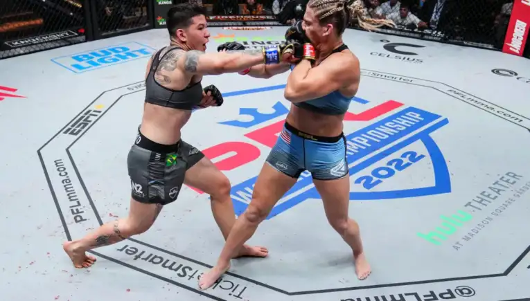 Two female mixed martial artists exchange punches inside an arena cage during a championship bout, centered on a mid‑ring exchange with the crowd watching above.