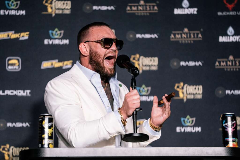 Man in a white suit speaks into a microphone at a press event, holding a cigar in one hand and wearing sunglasses.