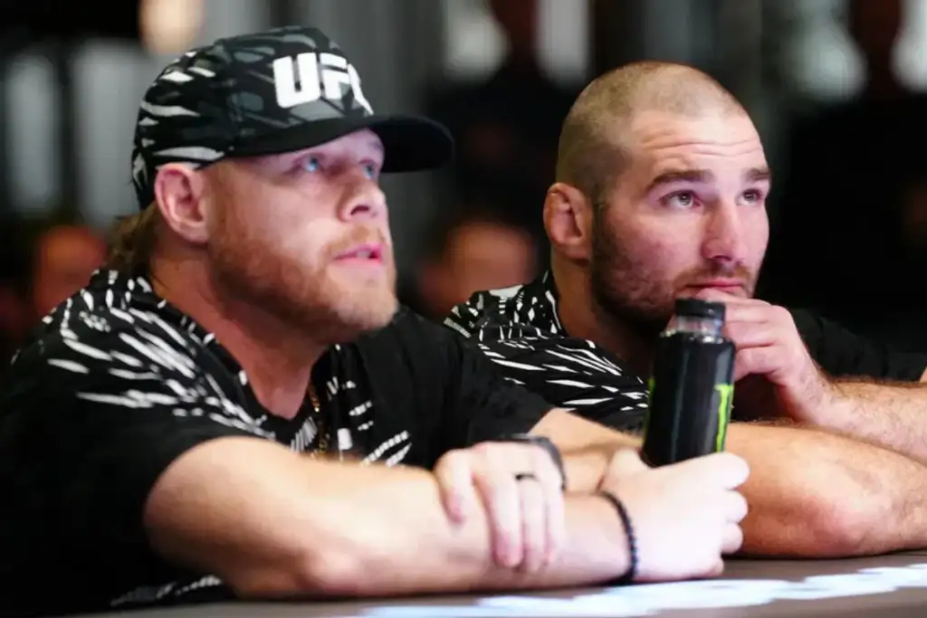Two men sit at a table, focused on something off-camera; one wears a UFC-cap and a black-and-white patterned shirt, the other rests his chin on his hand with a water bottle nearby.