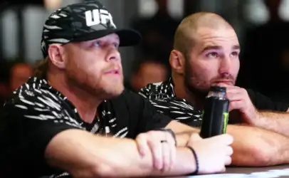 Two men sit at a table, focused on something off-camera; one wears a UFC-cap and a black-and-white patterned shirt, the other rests his chin on his hand with a water bottle nearby.