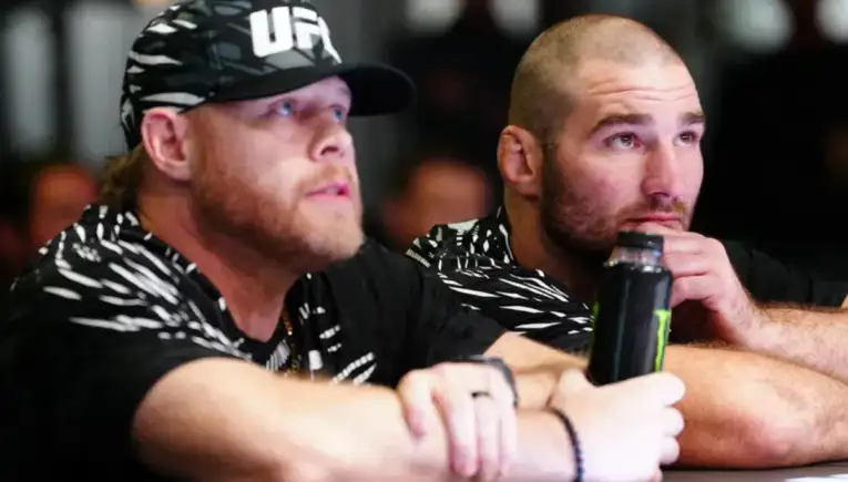 Two men sit at a table, focused on something off-camera; one wears a UFC-cap and a black-and-white patterned shirt, the other rests his chin on his hand with a water bottle nearby.