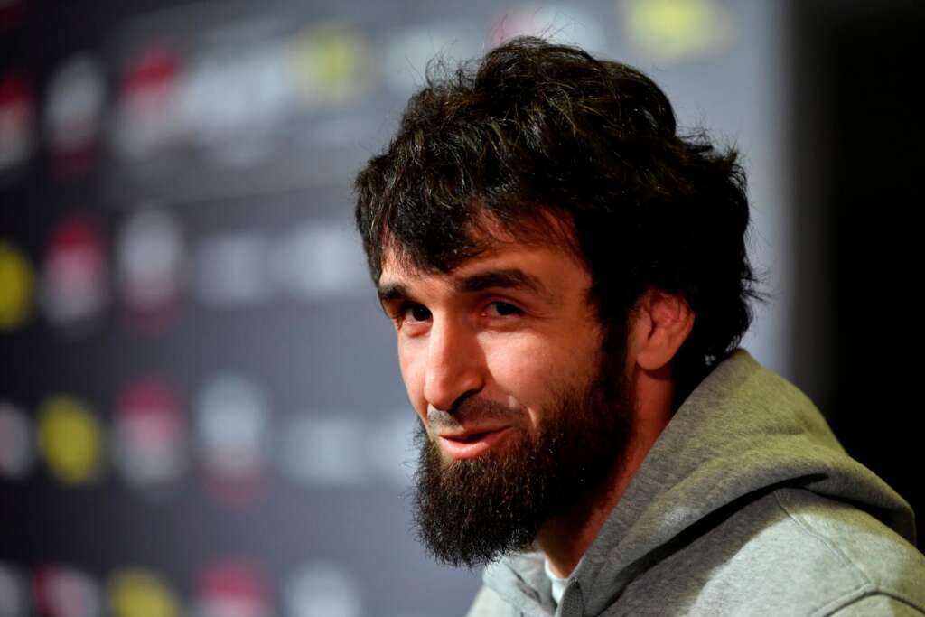 Bearded man in a gray hoodie, looking toward the camera with a soft, blurred backdrop behind him.