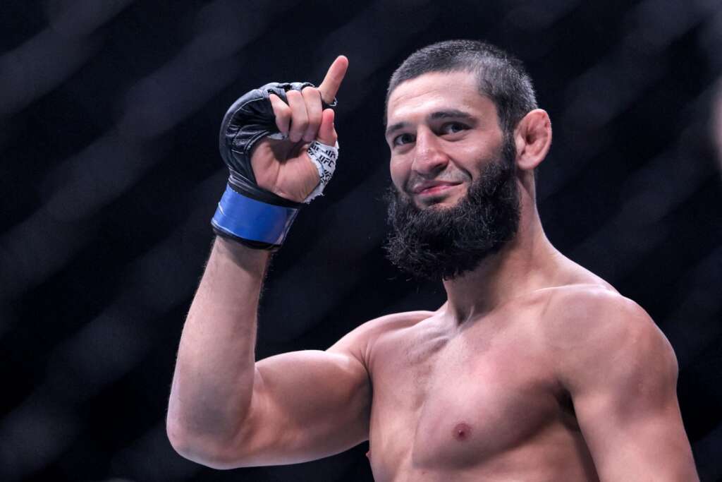 Bearded, shirtless male fighter raises his right fist in a victory pose inside an arena, wearing a taped hand wrap and glove.