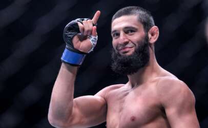 Bearded, shirtless male fighter raises his right fist in a victory pose inside an arena, wearing a taped hand wrap and glove.
