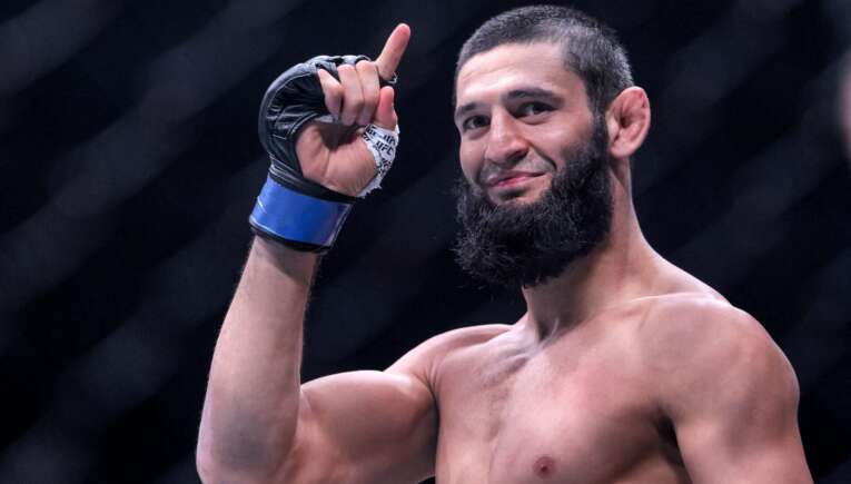 Bearded, shirtless male fighter raises his right fist in a victory pose inside an arena, wearing a taped hand wrap and glove.