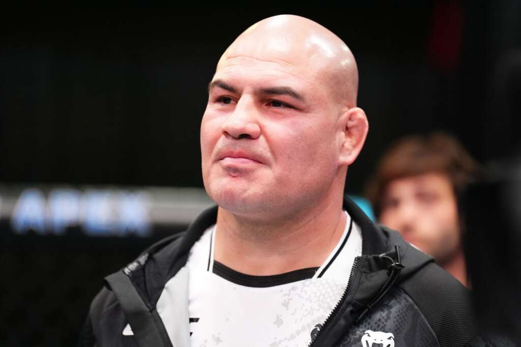 Close-up of a bald man in a black jacket, looking slightly to the left against a dark blurred background.