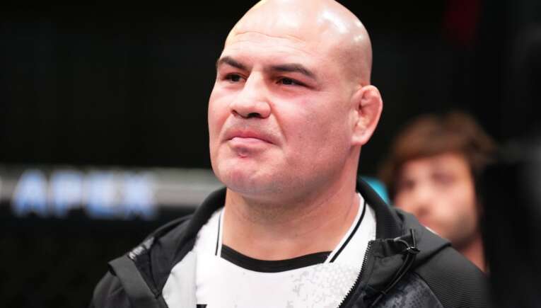 Close-up of a bald man in a black jacket, looking slightly to the left against a dark blurred background.