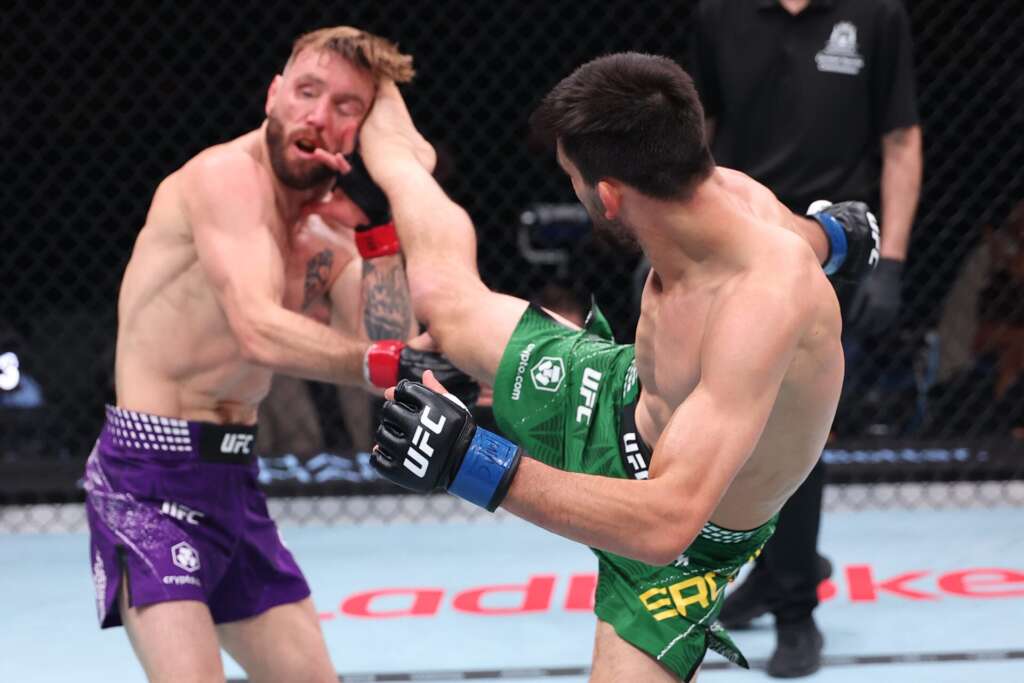 MMA fight inside a cage: fighter in green delivers a high kick to an opponent in purple shorts.