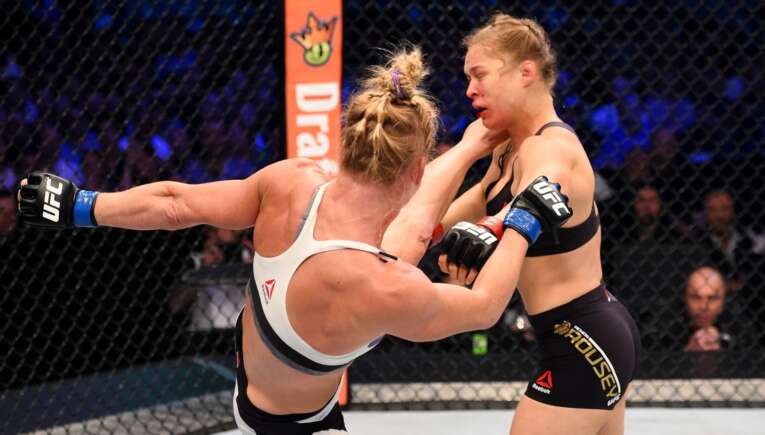 Two female UFC fighters in the octagon trading blows, one in a white top and the other in black shorts.
