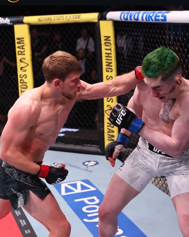 Two shirtless mixed martial artists exchange strikes inside a UFC cage; fighter on left throws a punch at a green-haired foe in white shorts, crowd watching behind.