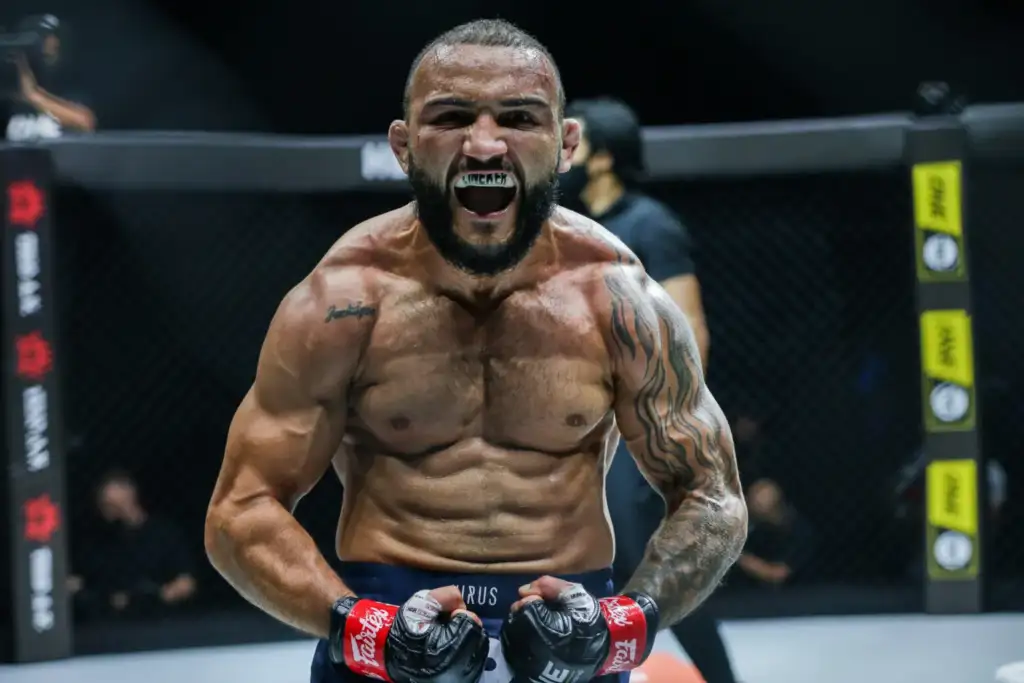 MMA fighter inside a cage, shouting with clenched fists, wearing red gloves and shorts, muscular and tattooed shoulders.