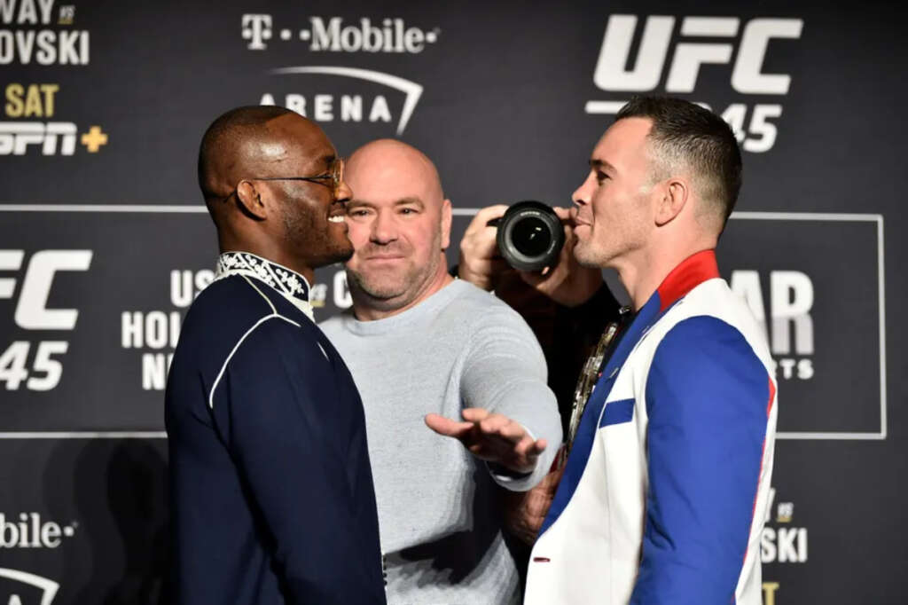 Two male fighters face off in a media event, a photographer between them aiming a lens toward the scene, with UFC branding in the background.