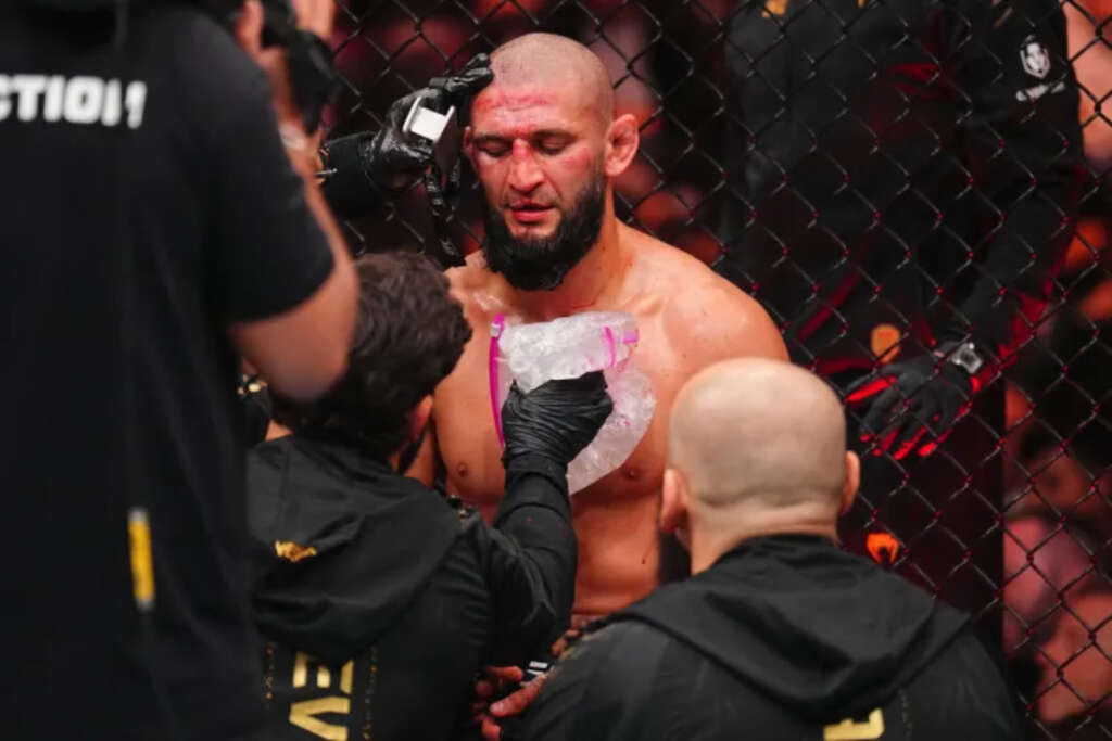 MMA fighter with a shaved head and beard receives medical treatment inside a cage, a team member placing a wrap on his chest.
