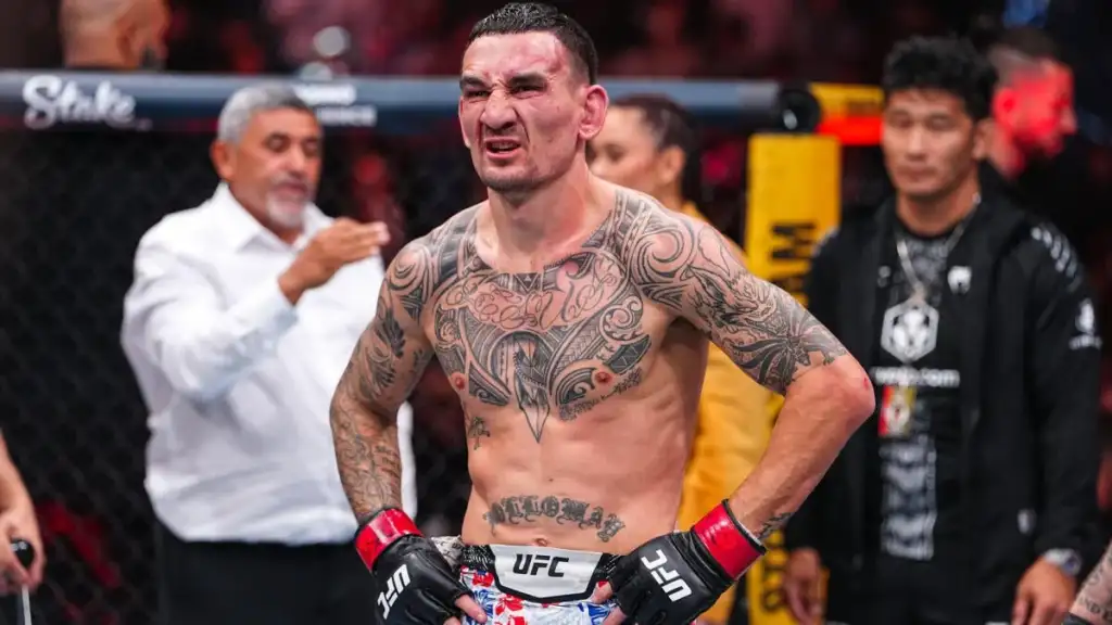 Tattooed UFC fighter with red gloves and hands on hips, grimacing inside a cage.Background shows officials and audience.