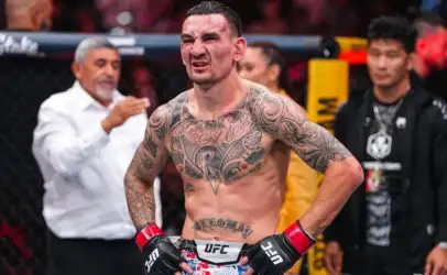 Tattooed UFC fighter with red gloves and hands on hips, grimacing inside a cage.Background shows officials and audience.