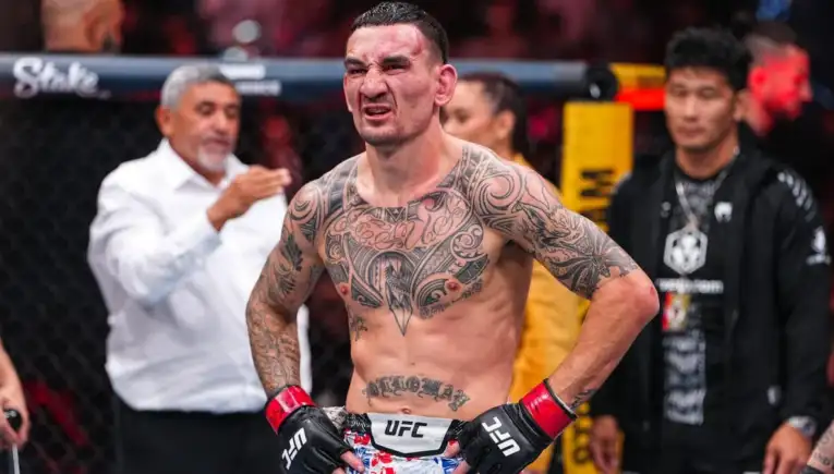 Tattooed UFC fighter with red gloves and hands on hips, grimacing inside a cage.Background shows officials and audience.