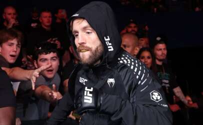 MMA fighter in a black hooded UFC jacket walks toward the camera as fans reach out from the crowd.
