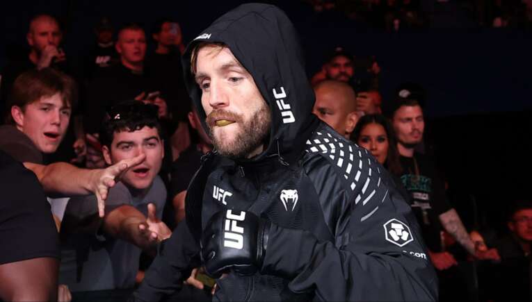 MMA fighter in a black hooded UFC jacket walks toward the camera as fans reach out from the crowd.