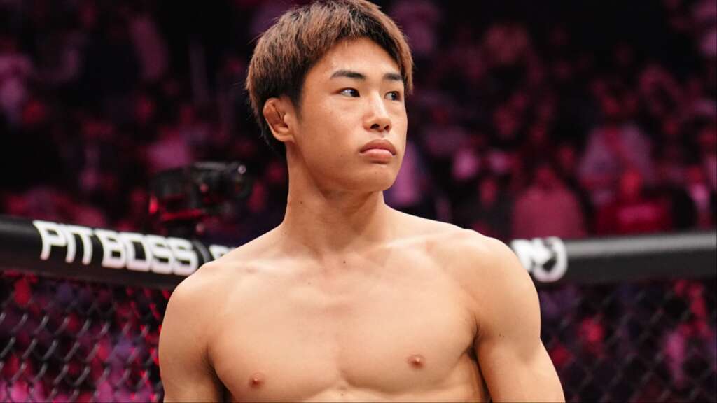 Shirtless male mixed martial artist in a cage, looking to the left with a blurred pink crowd behind.