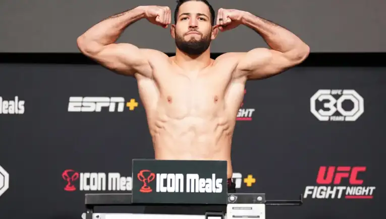 Male shirtless MMA fighter flexing on the weigh-in stage, arms raised, in front of branded backdrop (UFC, ICON Meals)