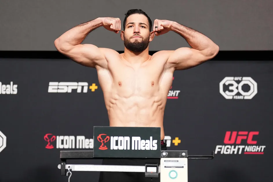 Male shirtless MMA fighter flexing on the weigh-in stage, arms raised, in front of branded backdrop (UFC, ICON Meals)