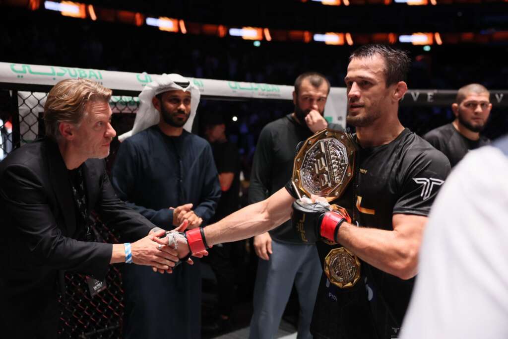 MMA fighter in black with a gold championship belt shaking hands with a coach inside the cage.