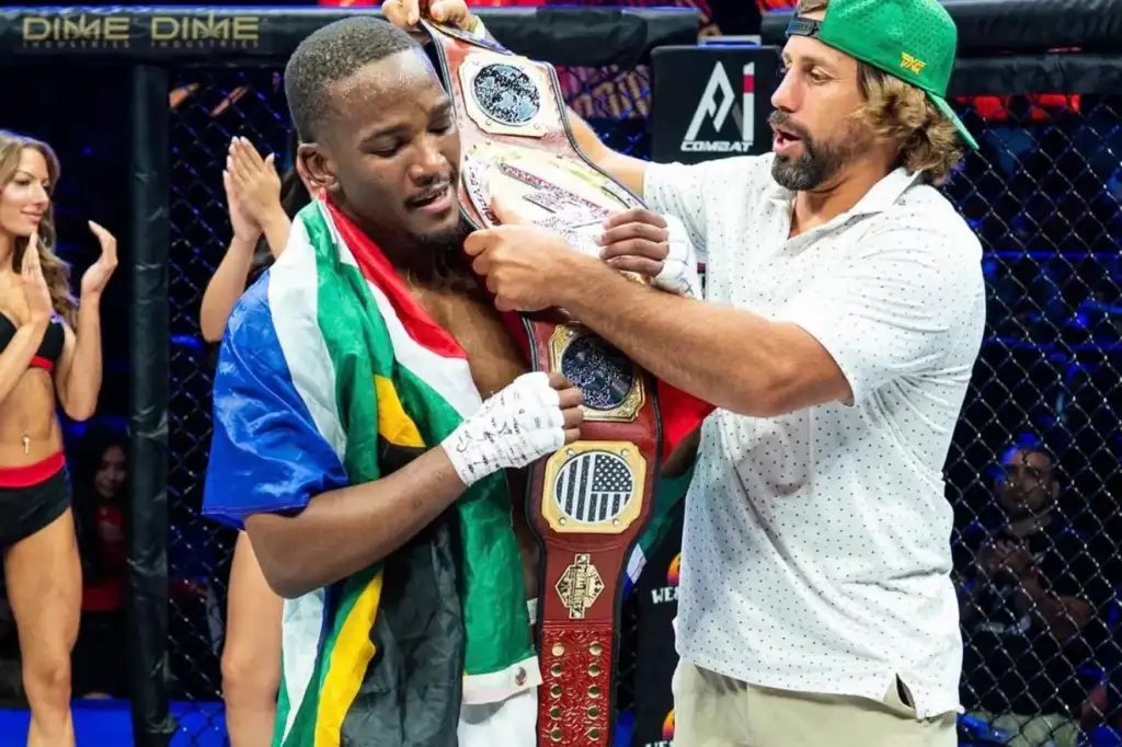 MMA fighter draped in a colorful flag, holding a championship belt as a coach adjusts it in the cage, audience applauding in the background.