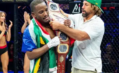 MMA fighter draped in a colorful flag, holding a championship belt as a coach adjusts it in the cage, audience applauding in the background.
