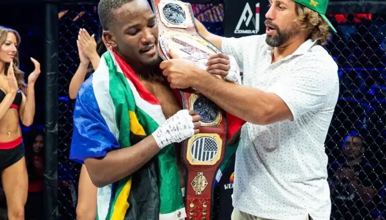 MMA fighter draped in a colorful flag, holding a championship belt as a coach adjusts it in the cage, audience applauding in the background.
