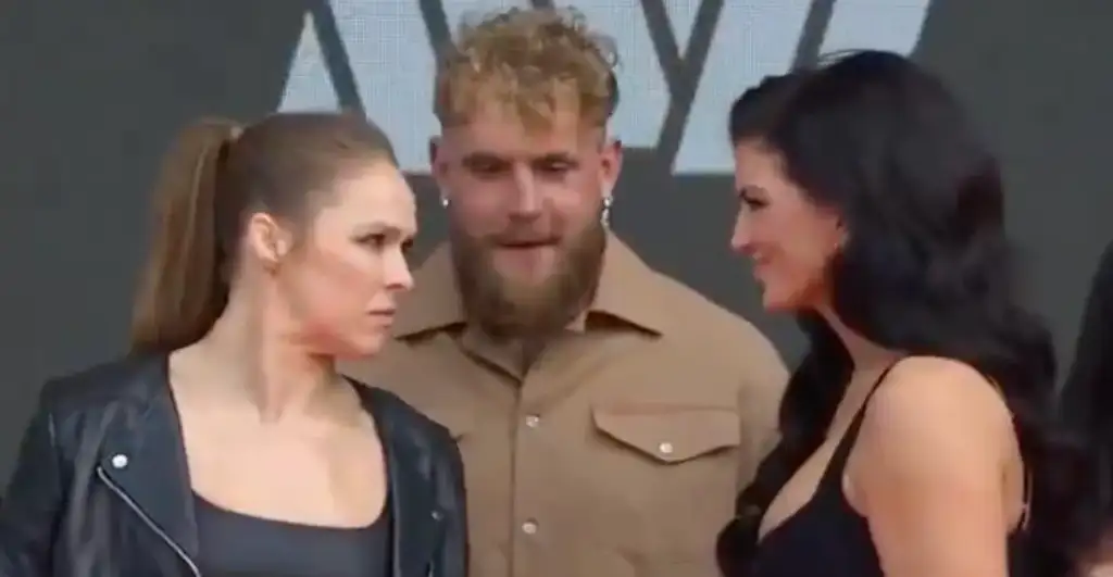 Three people stand on a stage: a woman in a black leather jacket on the left, a bearded man in a tan shirt in the middle, and a woman in a black dress on the right talking to him.