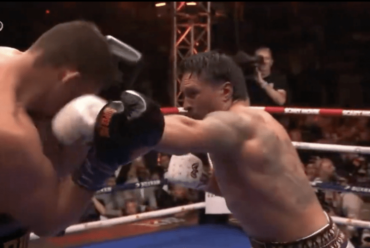 Two muscular boxers in a ring exchange punches, a glove making contact near the head as spectators watch from behind the ropes early in a bout.