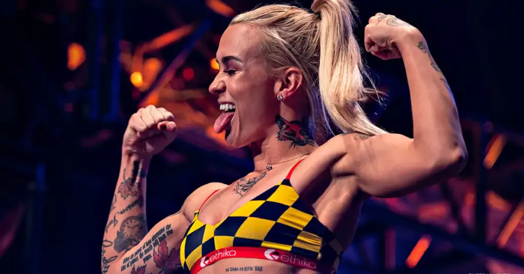 Female bodybuilder on stage flexing arms, tongue out, wearing a yellow and black checkered sports bra with visible tattoos on arms and neck.