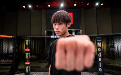 Young man in a black shirt extends a fist toward the camera inside a fenced fighting cage, focused on the lens in a dim arena setting.
