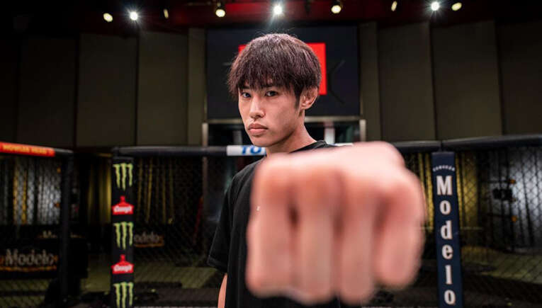 Young man in a black shirt extends a fist toward the camera inside a fenced fighting cage, focused on the lens in a dim arena setting.