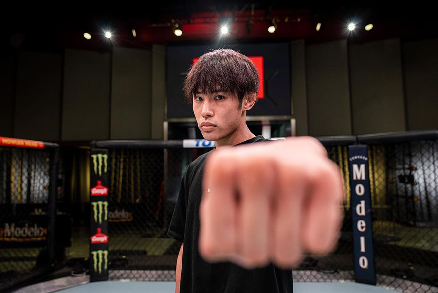 Young man in a black shirt extends a fist toward the camera inside a fenced fighting cage, focused on the lens in a dim arena setting.
