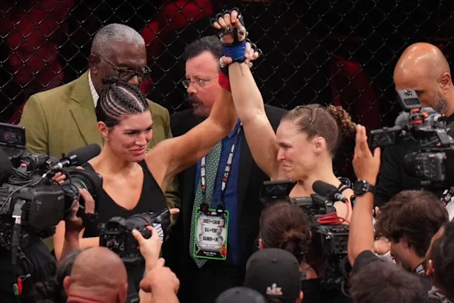 a526b0b4b6765cd12c953b7c765eb31c - AfroCombat Two female MMA fighters celebrate in the octagon as a referee raises the winner's arm, surrounded by photographers and a cheering crowd.