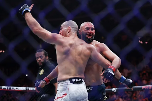 Two male UFC fighters clash in the octagon, one in white shorts landing a punch on the opponent in blue shorts, referee in the background.