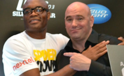 Two men pose together at an event, one in a white T-shirt with logos and the other in a black shirt, smiling with their arms around each other.