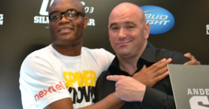 Two men pose together at an event, one in a white T-shirt with logos and the other in a black shirt, smiling with their arms around each other.